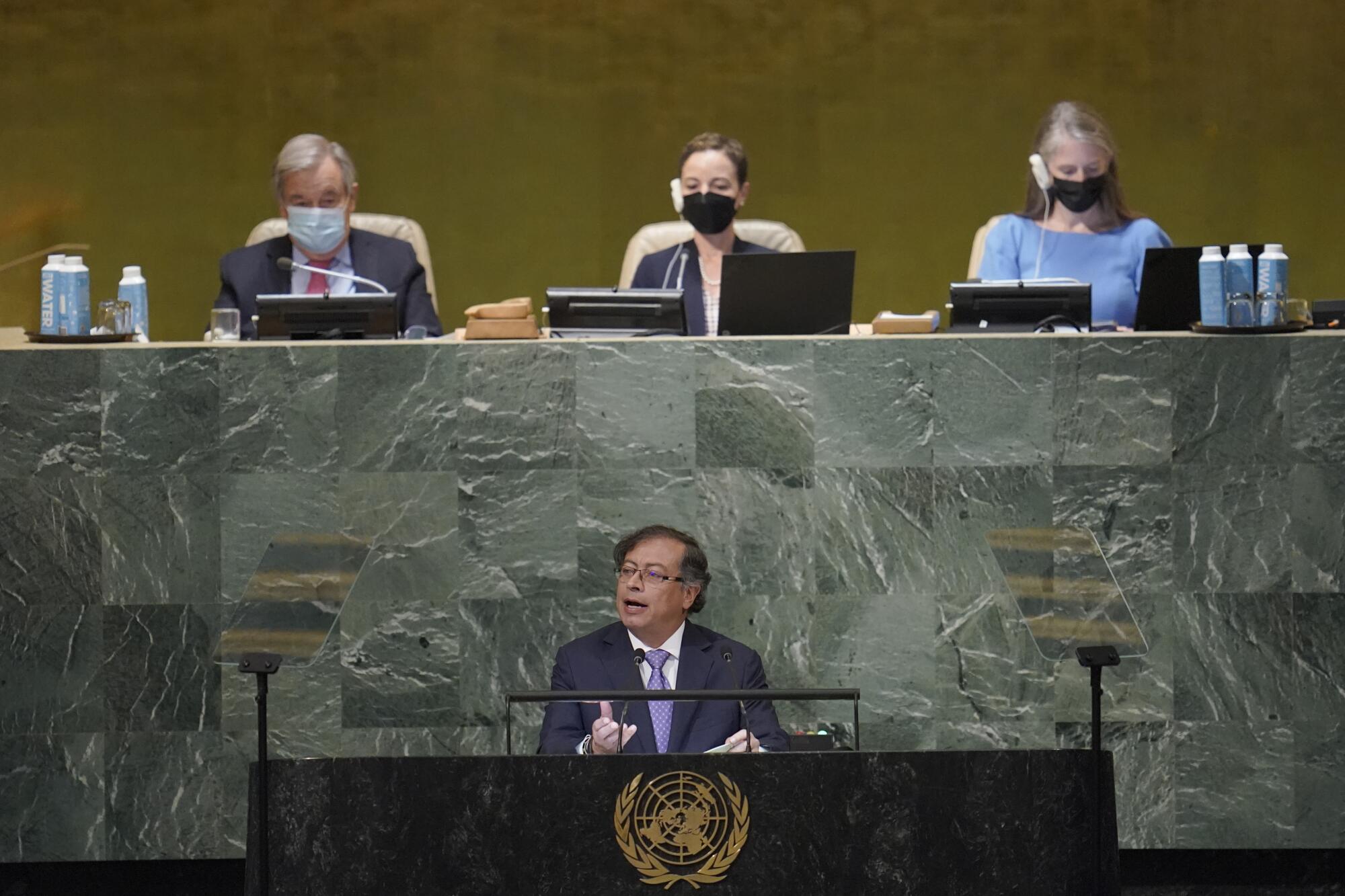 A man speaks at a lectern while three people sit behind and above him.