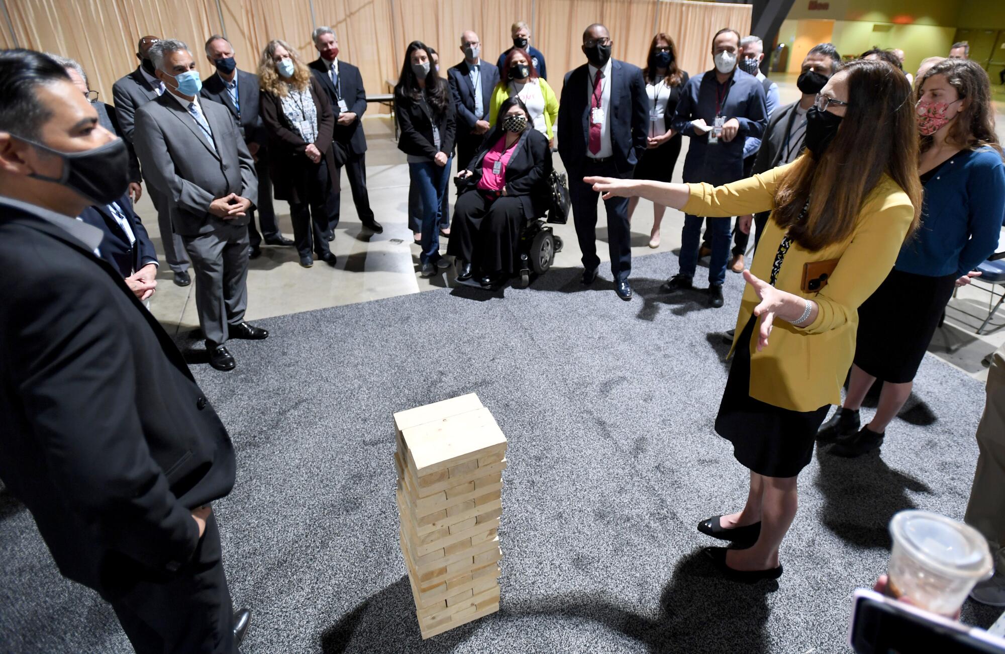 A woman speaks to a crowd of officials next to a large set of Jenga blocks