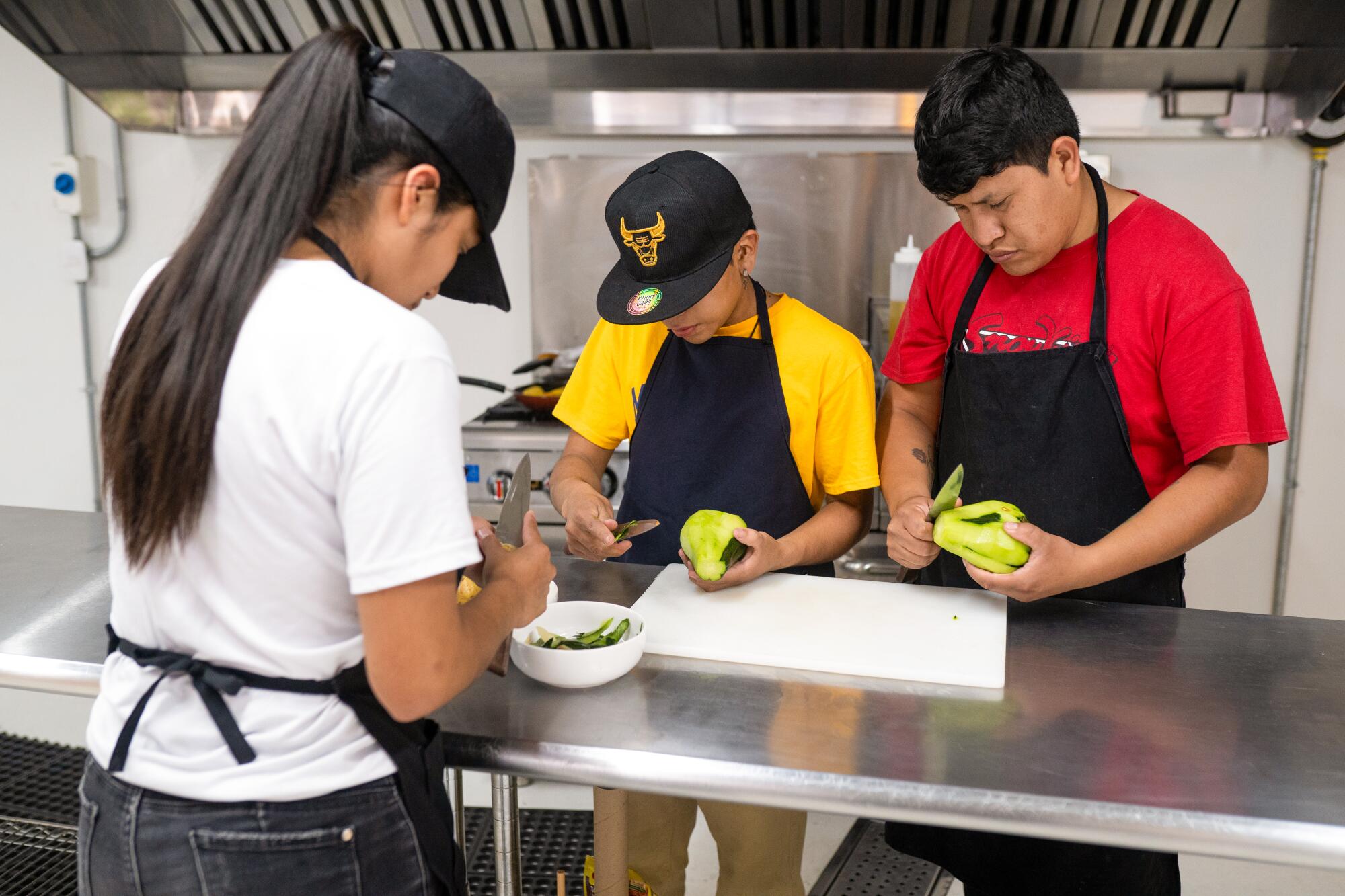 Three young cooking students peel a green squash standing at a metal table.