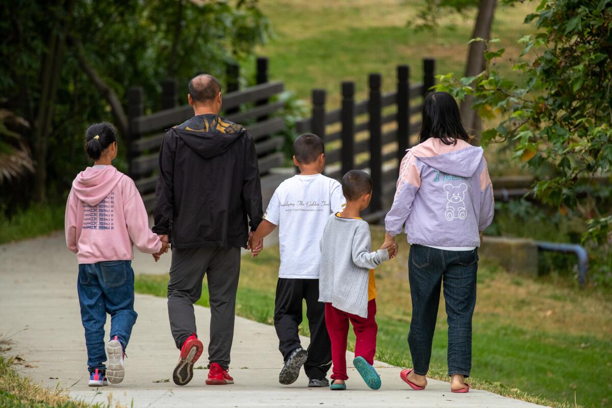 Two adults and three children walk on a park path, away from the camera while holding hands