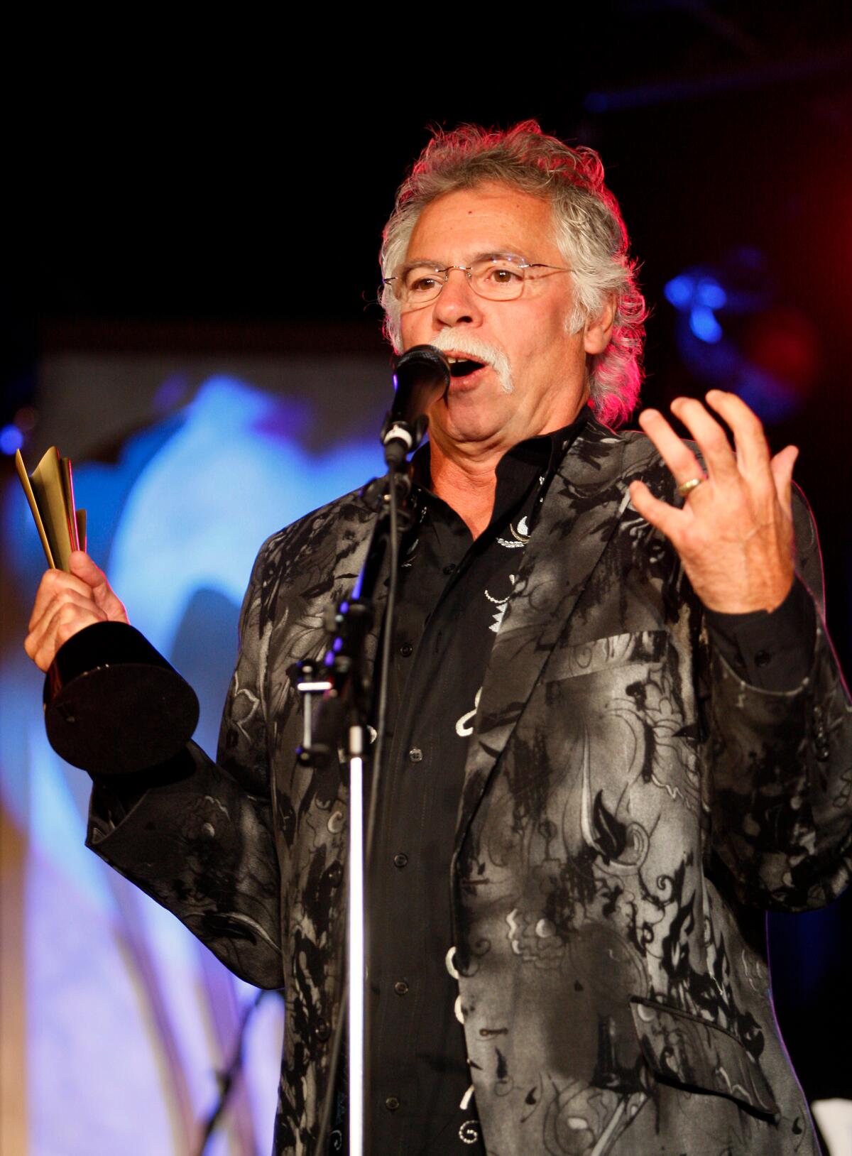 Joe Bonsall holds a gold and black trophy and talks into a mic while wearing a black suit jacket with floral designs