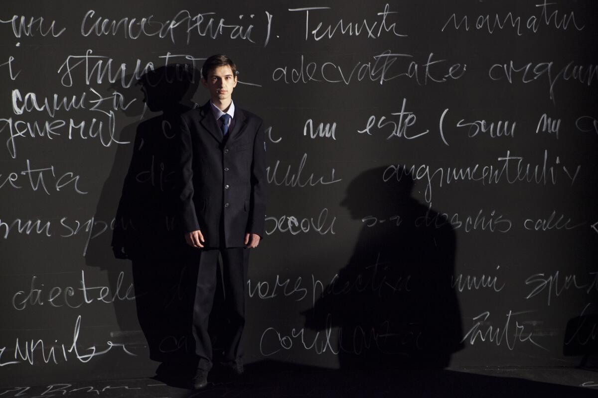 An adolescent stand in front of a chalkboard wall of text in the movie “Uppercase Print.”