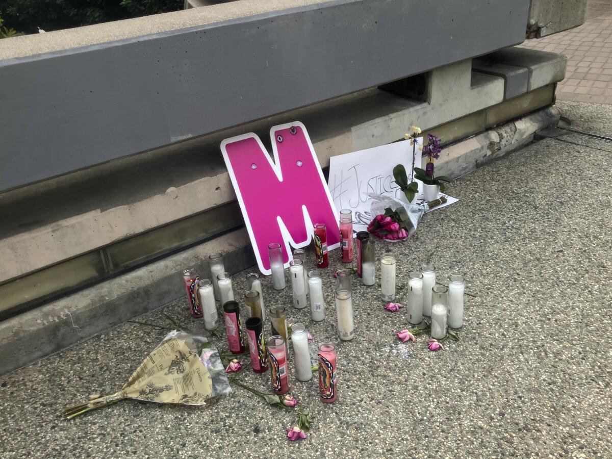 A pink letter M propped up against a barrier next to flowers, notes and over 20 candles arranged on pavement.