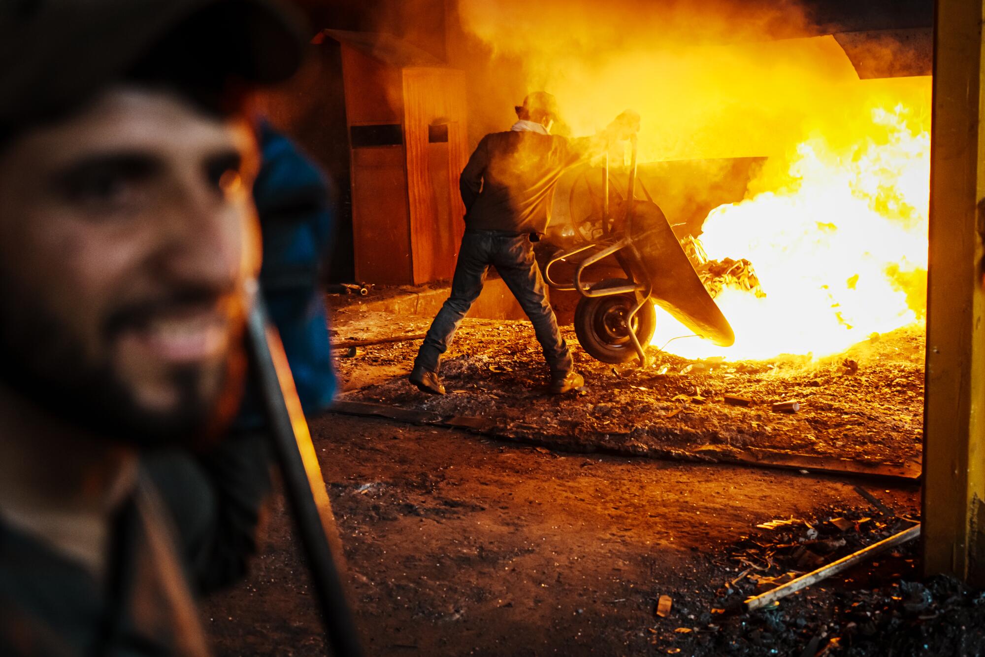 A man tips a wheelbarrow's contents into flames in a furnace