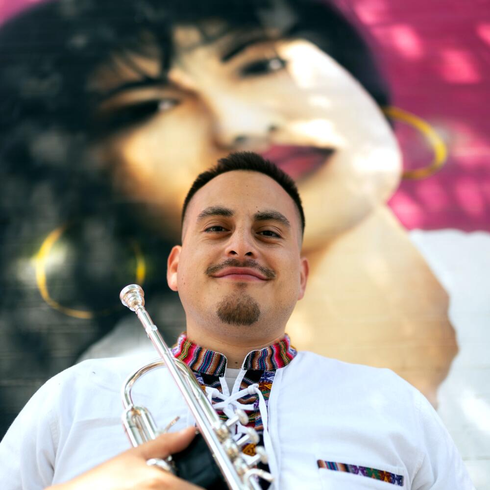 Holding a trumpet, Daniel Flores poses in front of a Selena Tribute mural in Chicago
