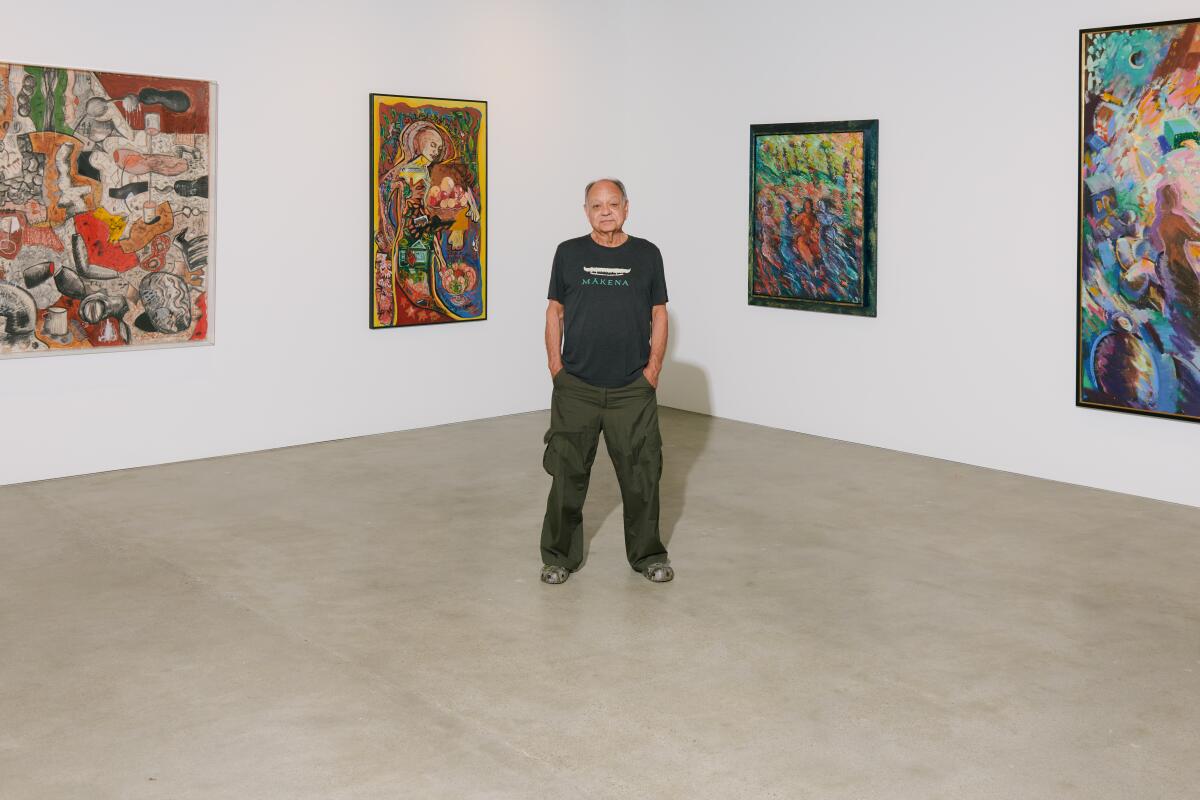 A man wearing a T-shirt and cargo pants stands among paintings in a gallery.