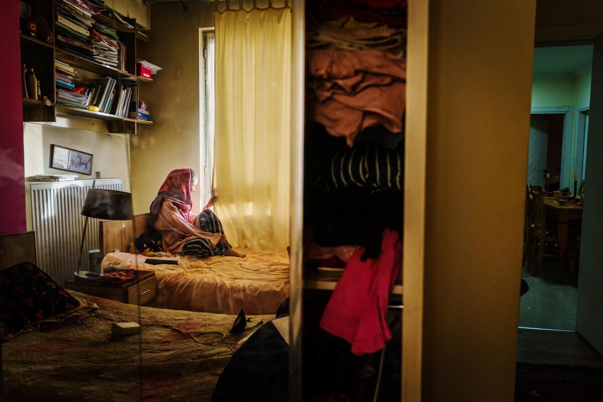 A young woman in a head covering sits in her bedroom on her bed and looks out the window