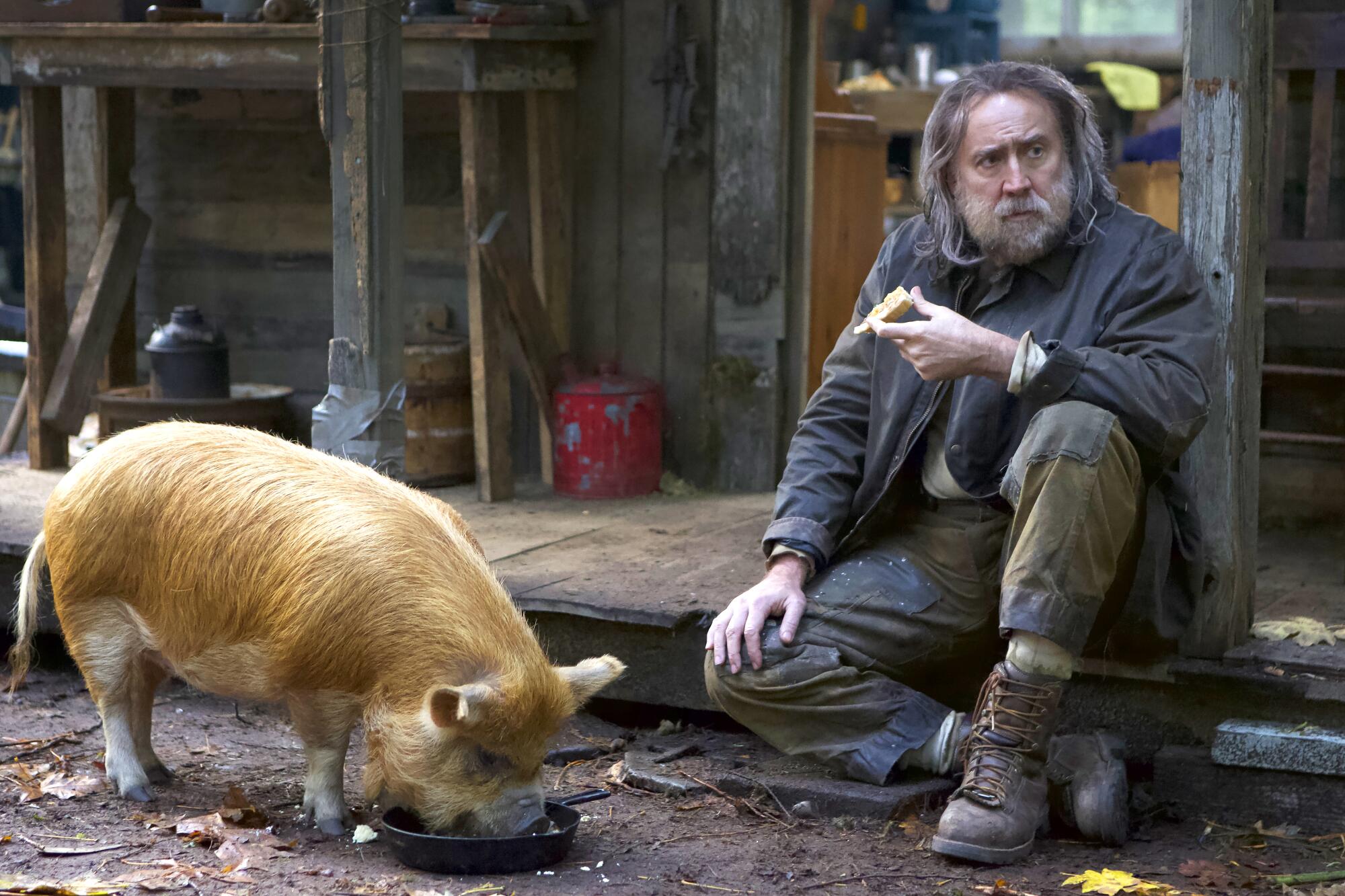 A long-haired and bearded Nicolas Cage sits down outside to eat while his pet pig chows down in a scene from "Pig." 