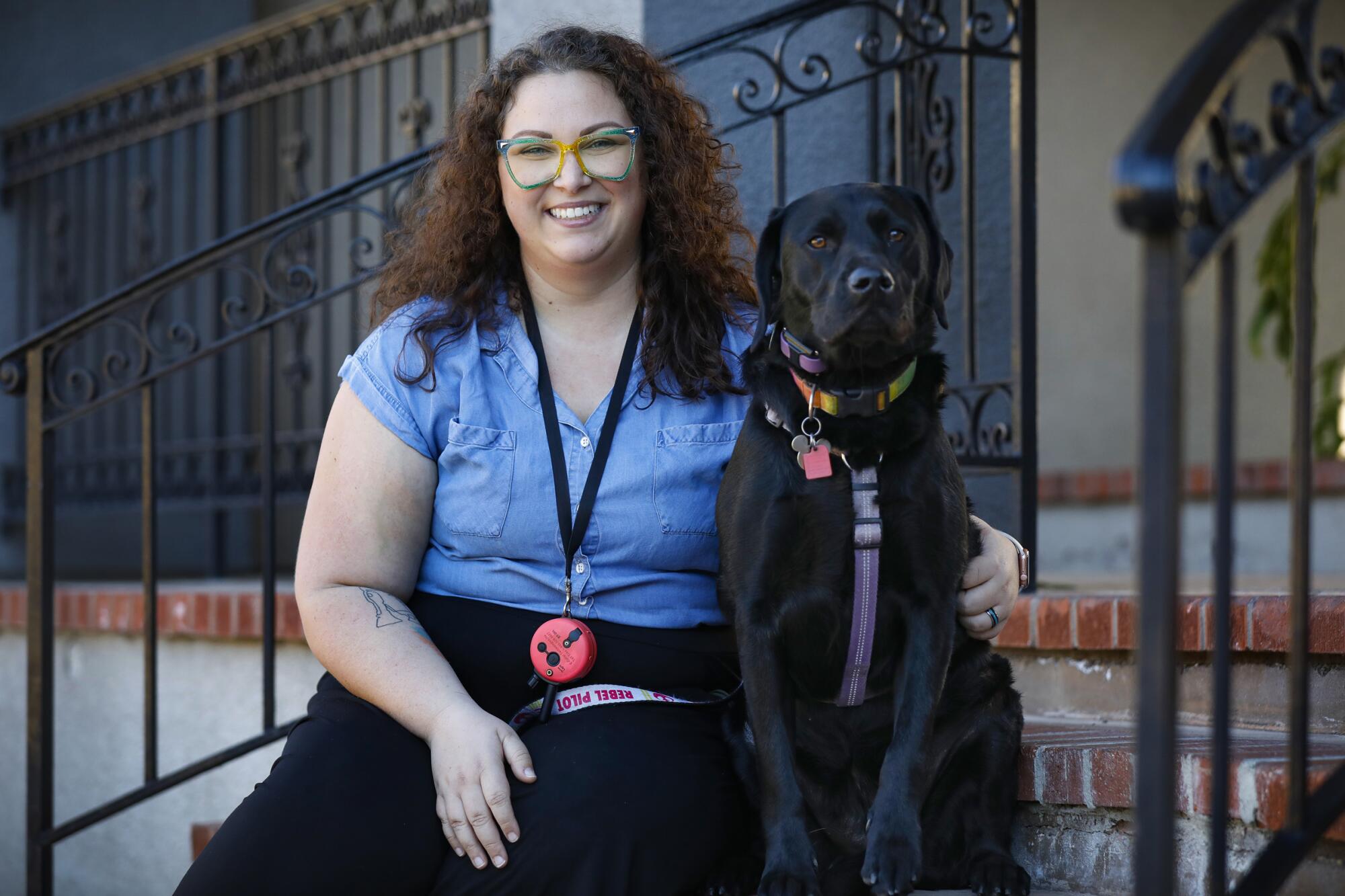 a woman wearing glasses sits on an apartment stoop with her service dog, a black lab