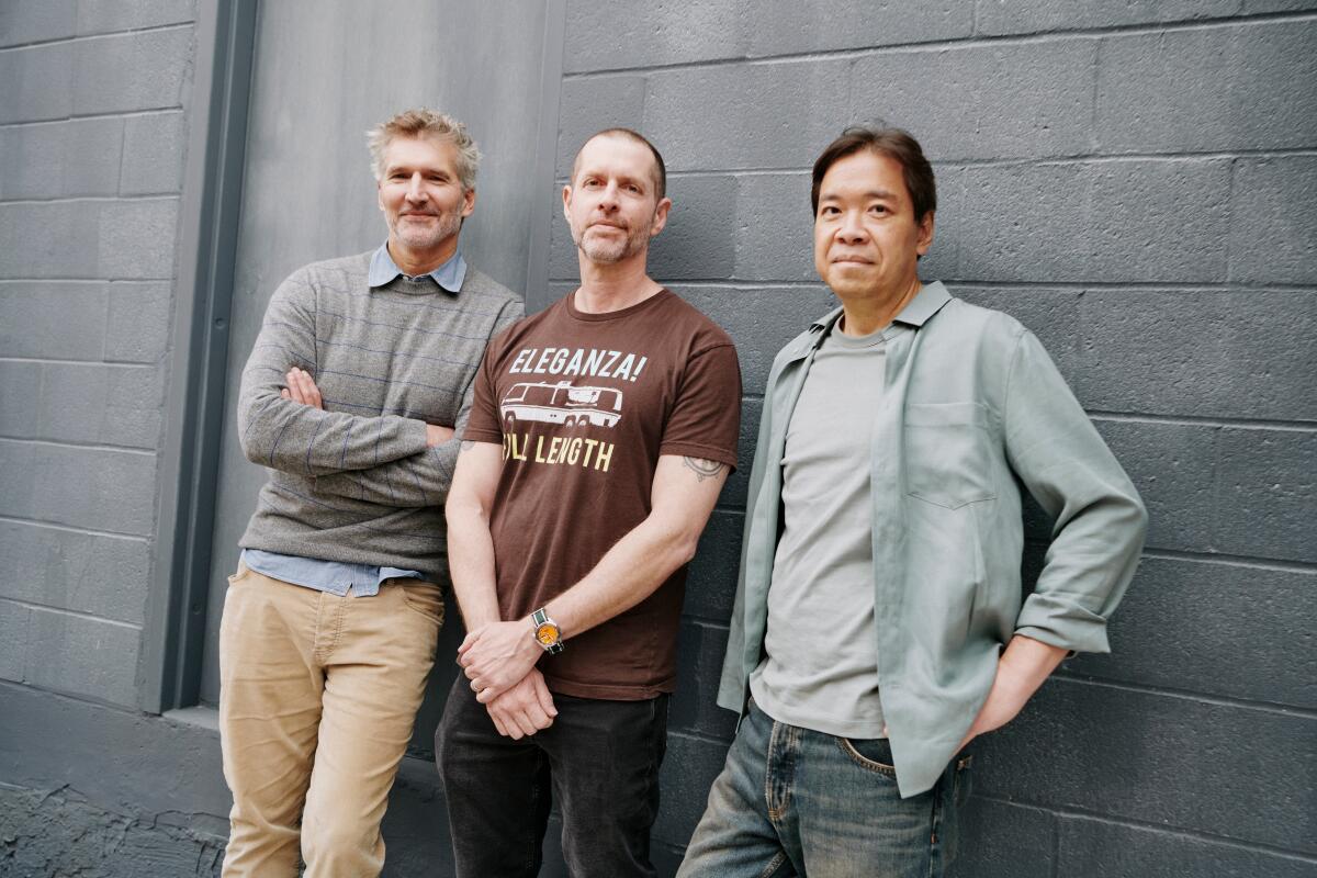 Three men dressed casually lean against a cement-block wall for a portrait.
