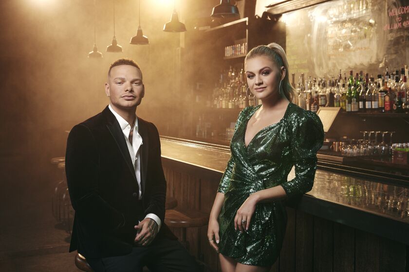 “CMT Music Awards” cohosts Kane Brown and Kelsea Ballerini pose by the counter in a hazy bar.