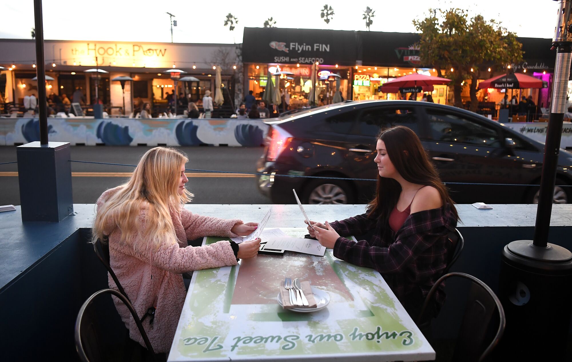 Two women, unmasked, speak across an outdoor table at a restaurant.