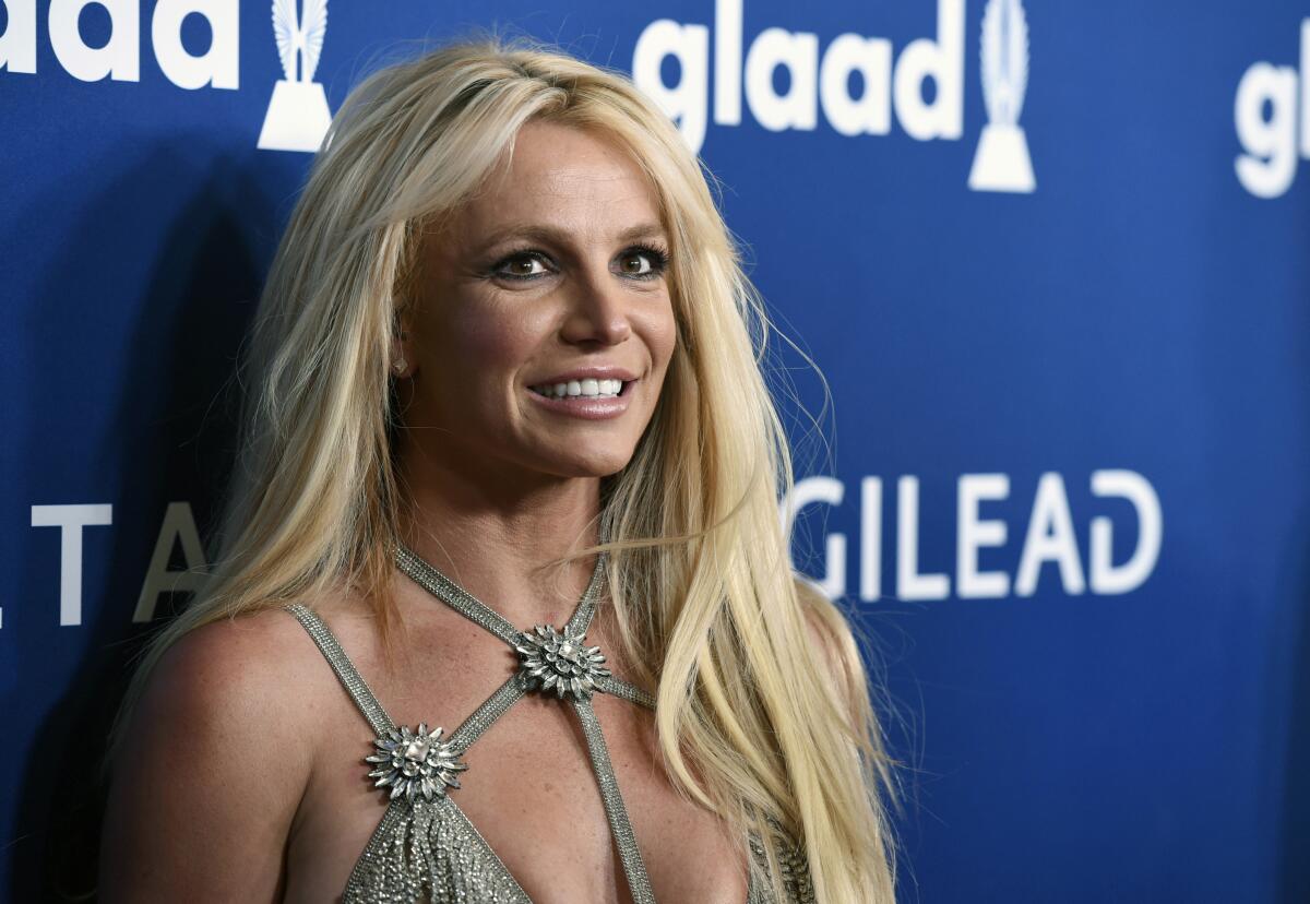 Britney Spears smiles in a sparkling, strappy silver gown while posing against a dark blue background