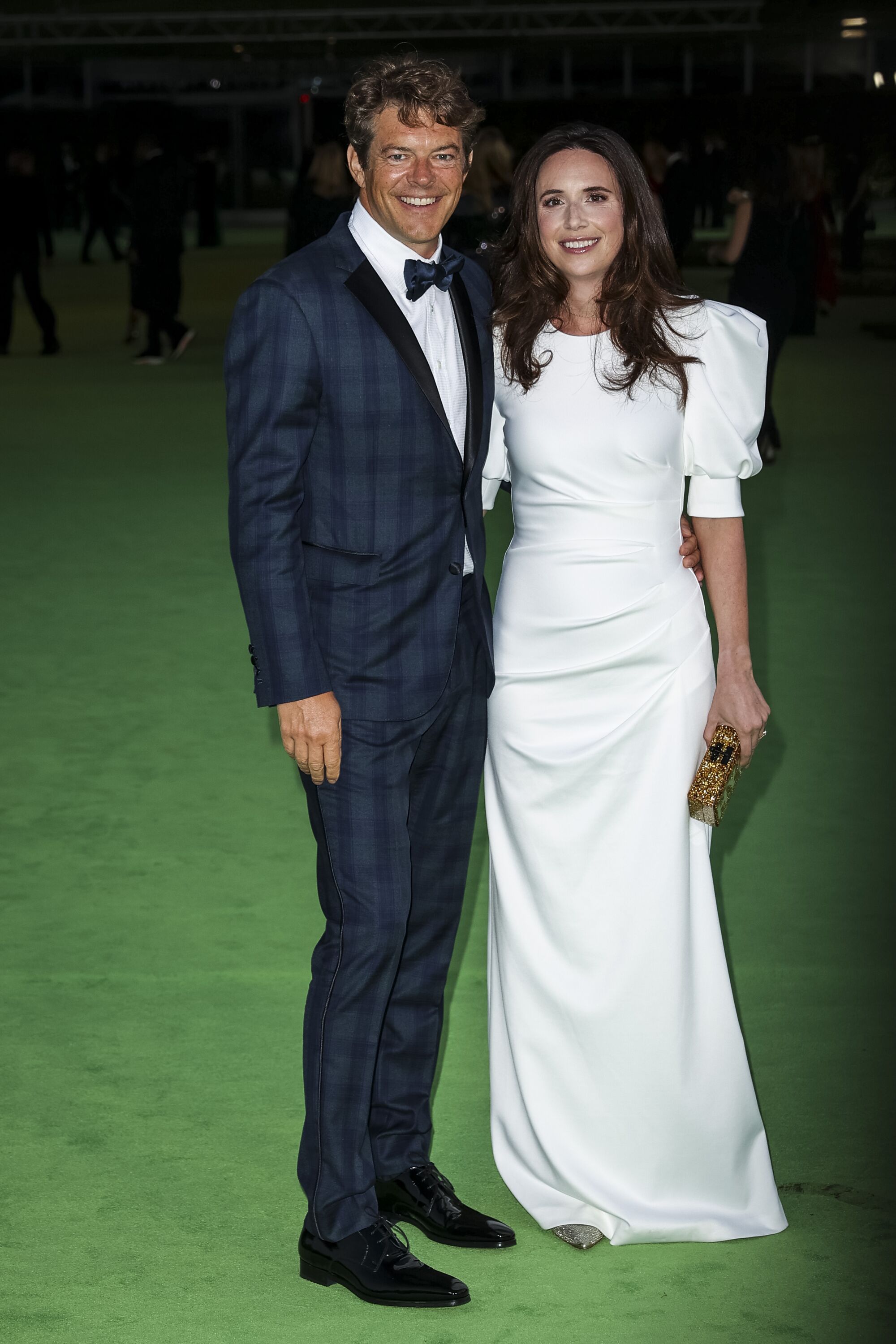 A man in a plaid, blue tuxedo and a woman in a white dress posing on a green carpet
