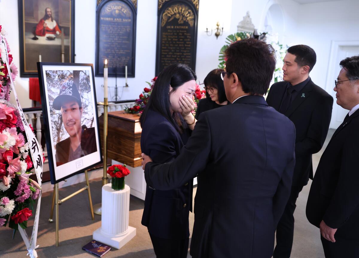 A woman is consoled by others as she cries in front of a large framed photo of a man