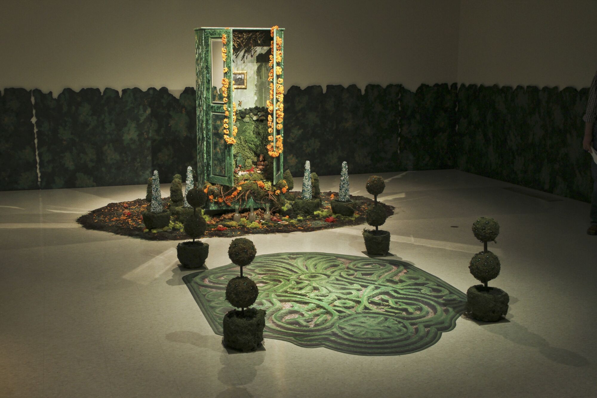 An armoire, painted green and strung with flowers, is presented in a dramatically lit gallery surrounded by plants.