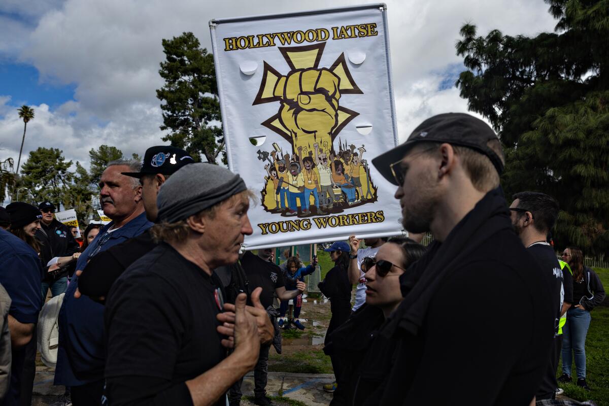 A trio of people talking at a rally in a public park. One is holding a 'Hollywood IATSE' sign with a golden fist.