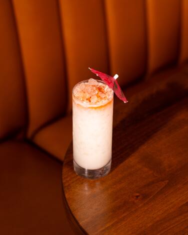 A milky-colored cocktail in a tall glass with an orange rim and paper umbrella