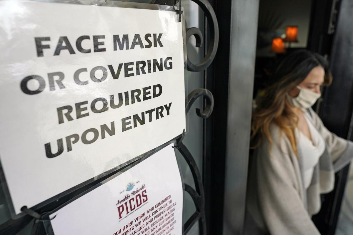 A woman in a surgical mask next to a sign that says "Face mask or covering required upon entry"