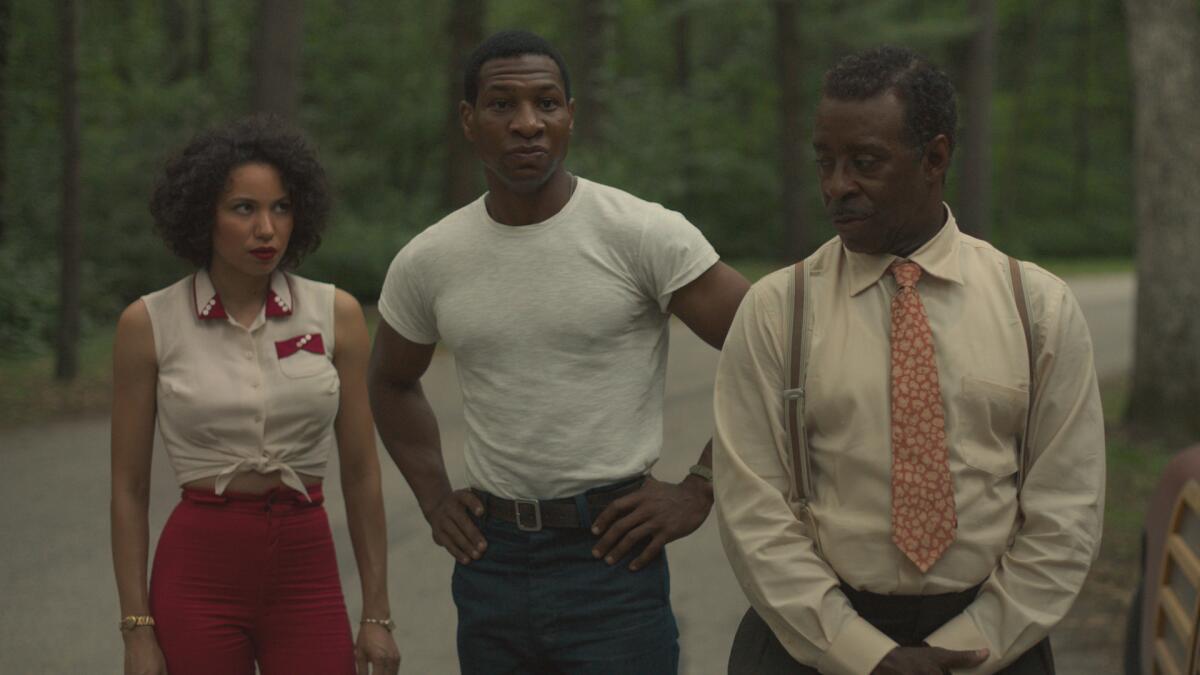 A woman and two men stand on a road beside a wooded area.