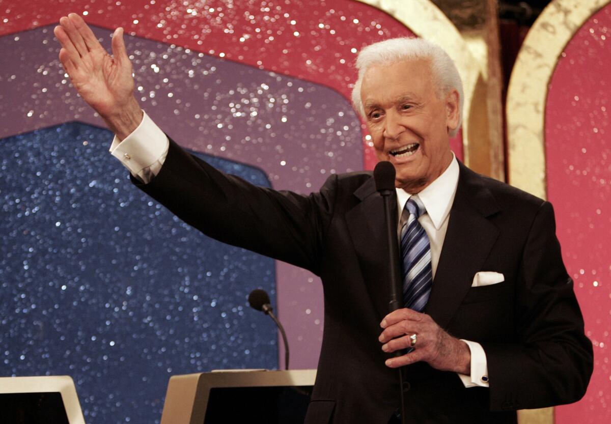Bob Barker, in a dark suit, gestures broadly with one hand while speaking into a microphone on a game show set.