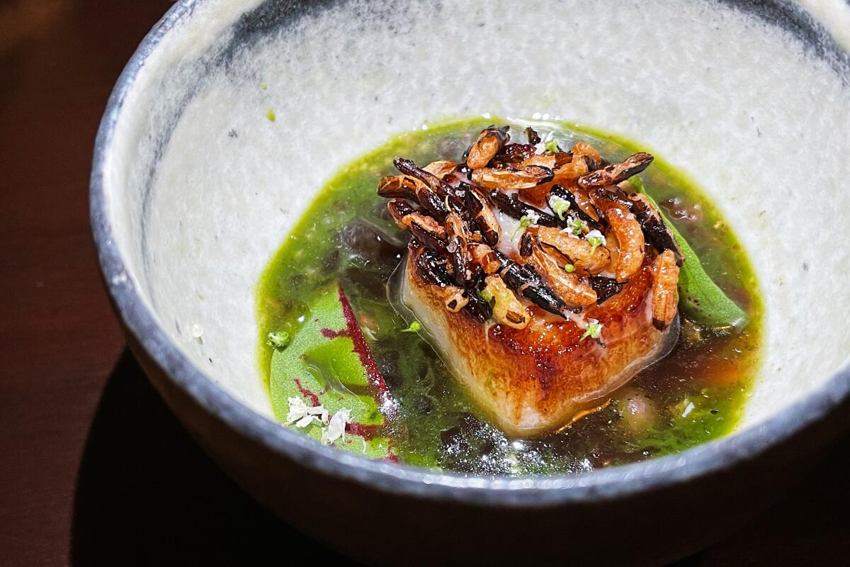 A browned Hokkaido scallop topped with puffed rice and sitting in a green broth in ceramic bowl at Baroo Los Angeles