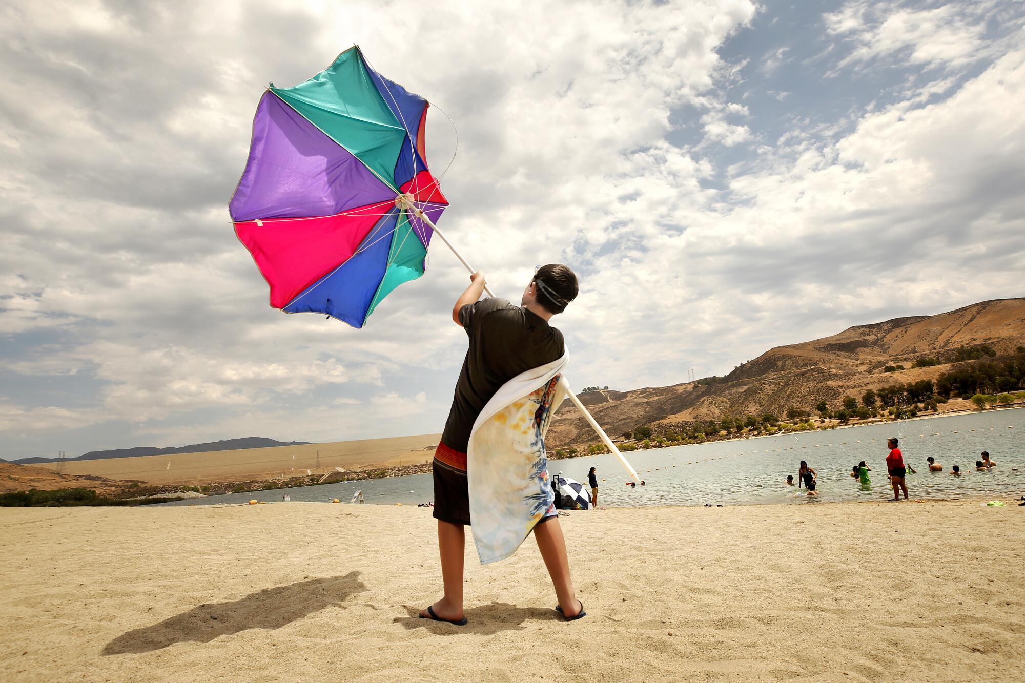 A boy on a sandy lakeshore holds an umbrella turned inside out by wind