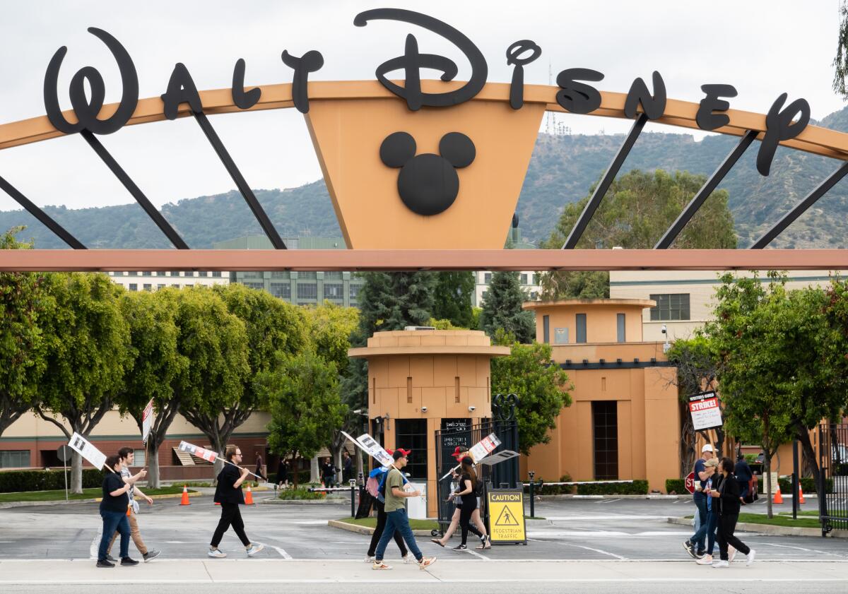 Picketers walk before a sign reading "Walt Disney," with a Mickey Mouse symbol underneath.