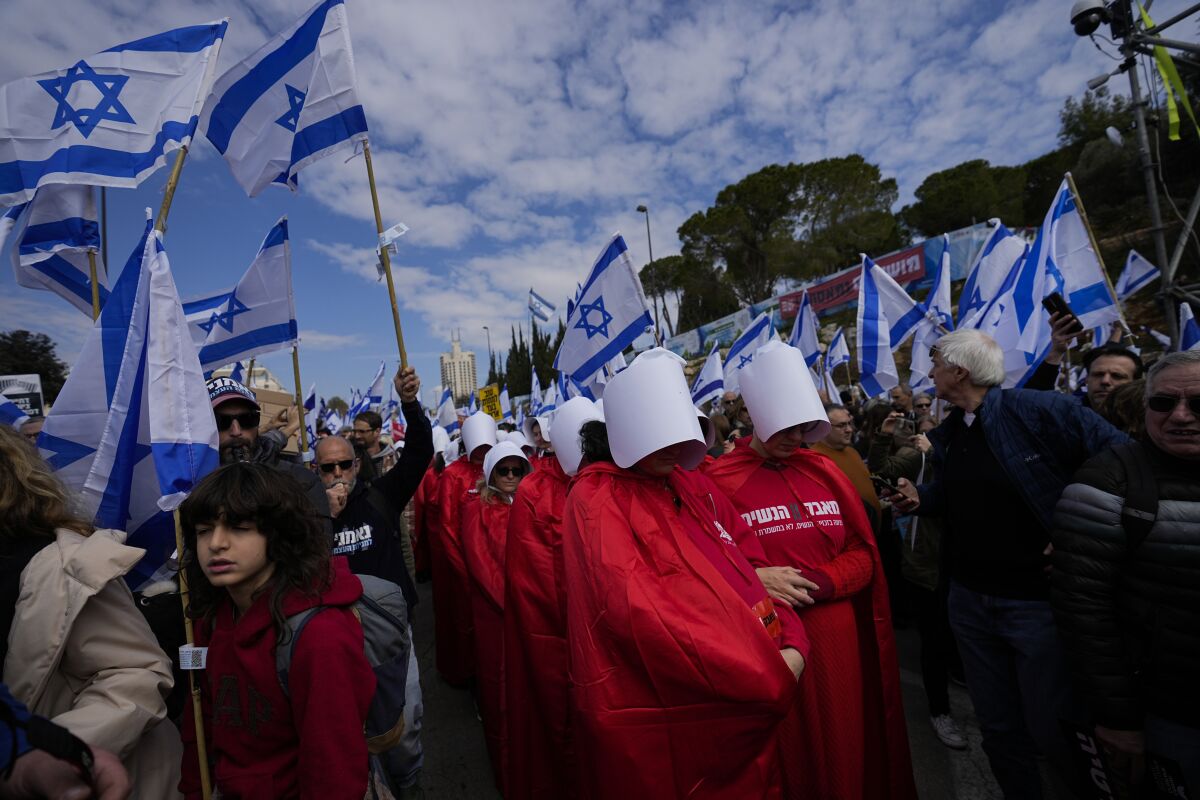 Israelis with flags or in red dresses and white caps protest outdoors