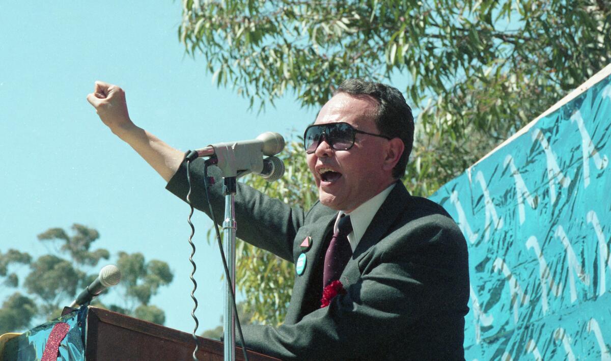A man in a suit, sunglasses and pink triangle pin signifying gay pride speaks at a microphone on a dais, his fist in the air.