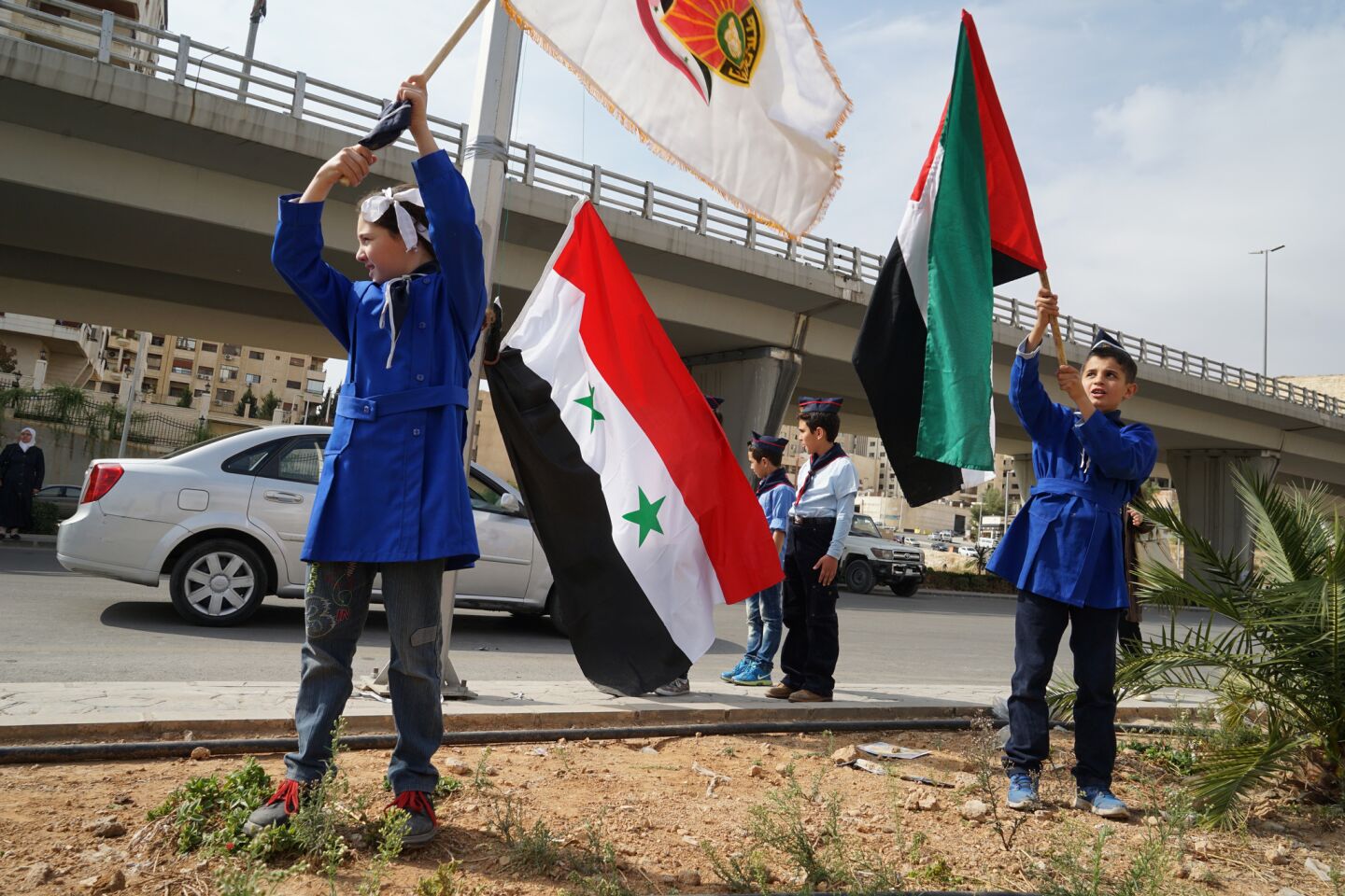 Elementary school children in a ruling party youth group wave Syrian flags along a street.