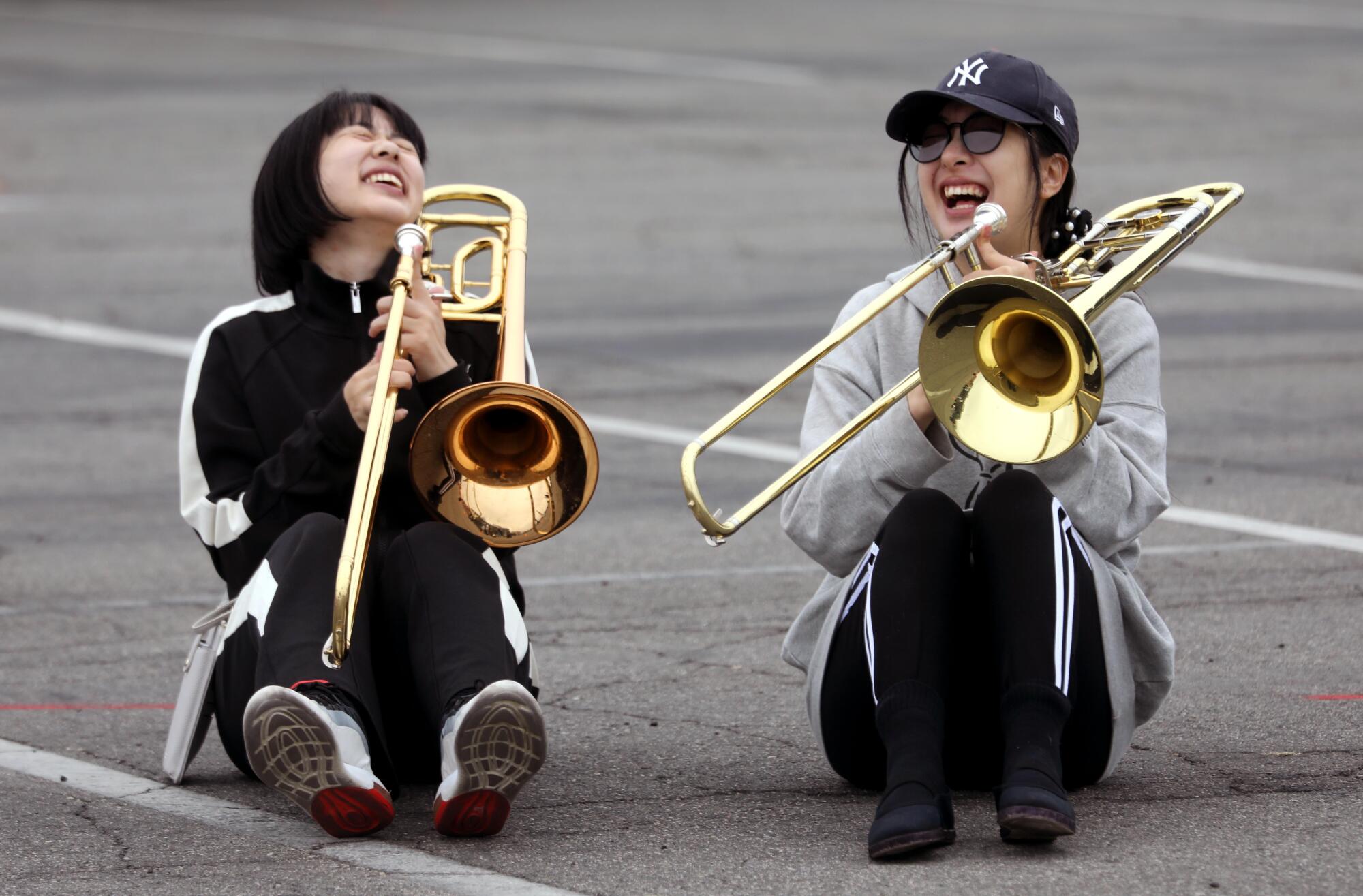 Two girls with trombones laugh while sitting in a parking lot.