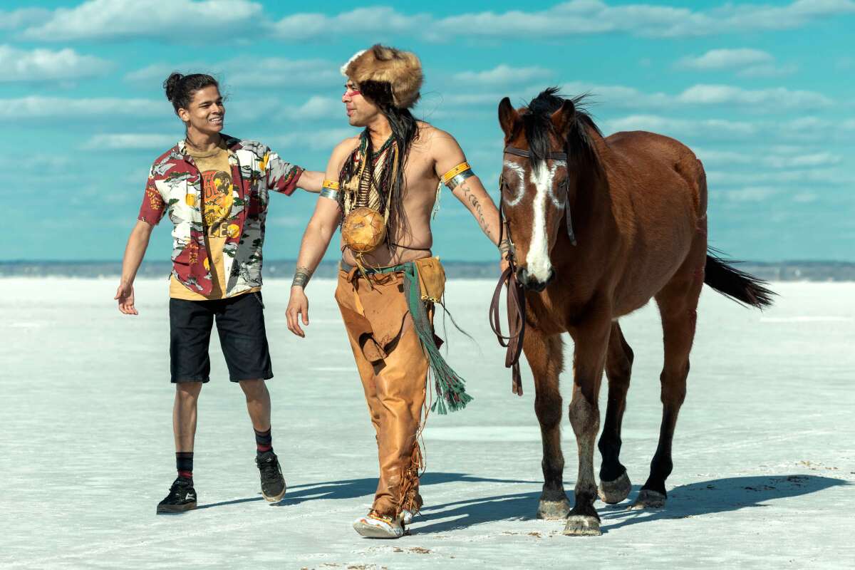 A teenage boy walking next to a man in a Native American loincloth leading a brown horse.
