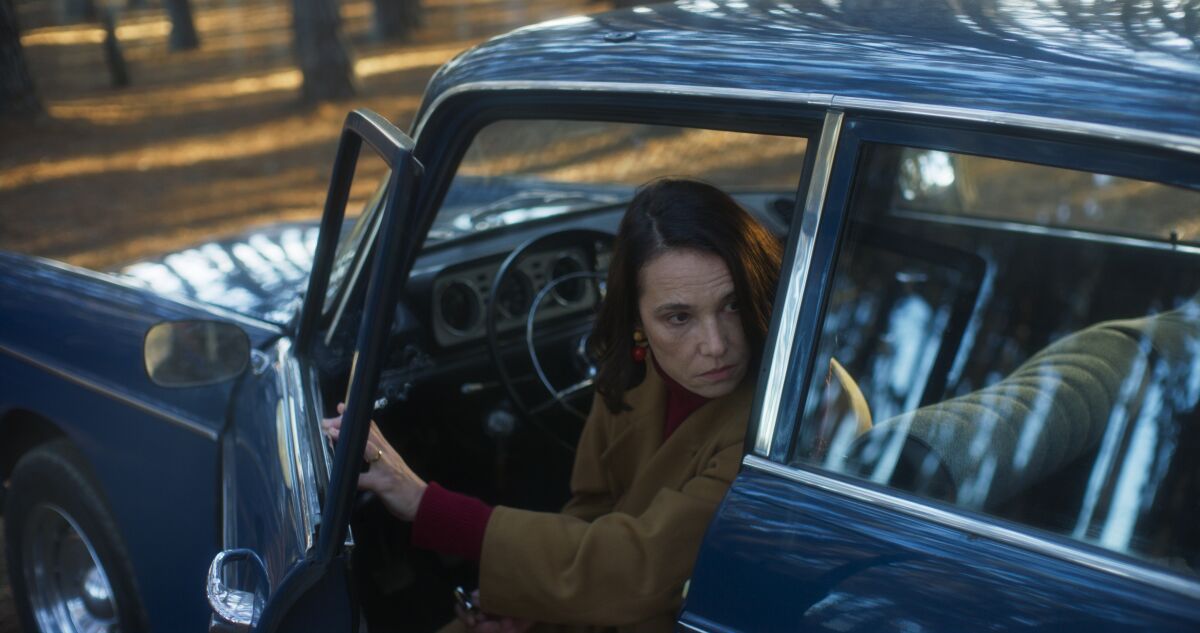 A woman seated in a car opens the door to look behind her.