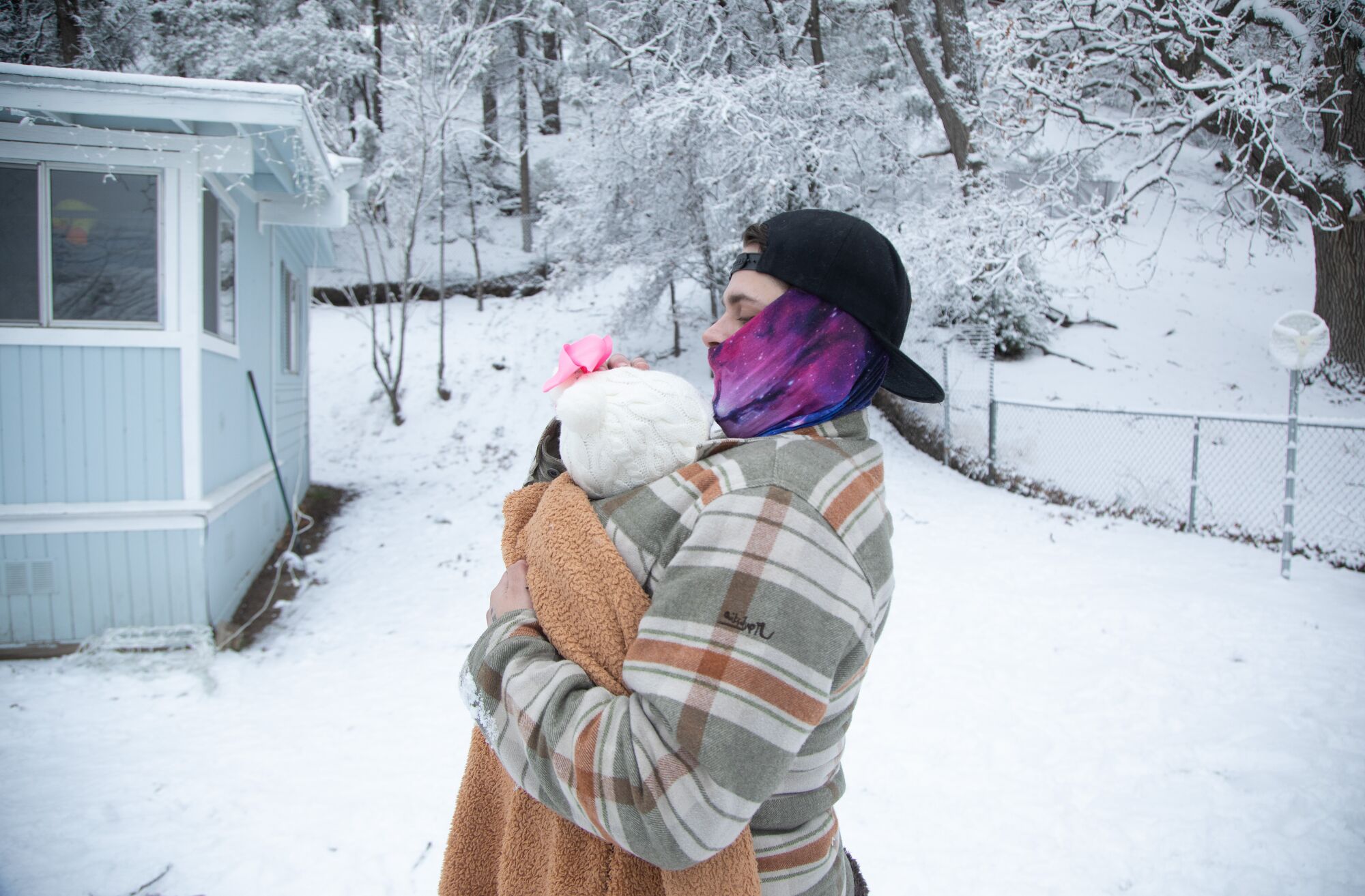 A man carries a baby, both bundled up against the cold, in a snowy yard