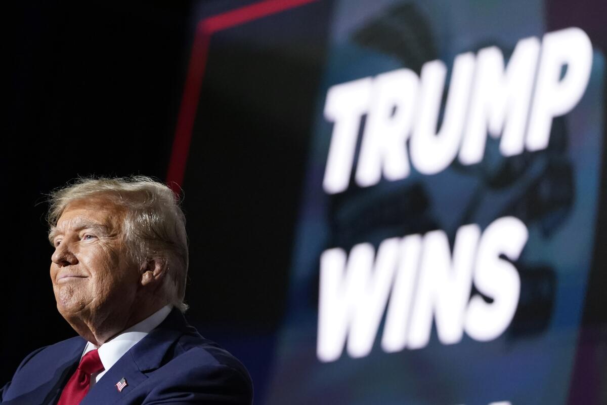 Former President Donald Trump speaks in front of a sign reading "Trump Wins"