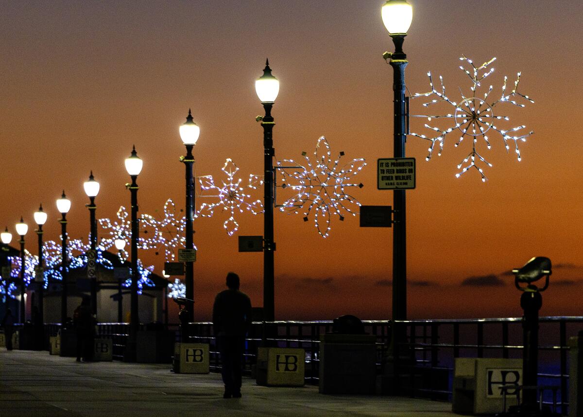 Lights shaped like big snowflakes hang above Huntington Beach Pier as pier walkers are silhouetted by the sunset's glow.