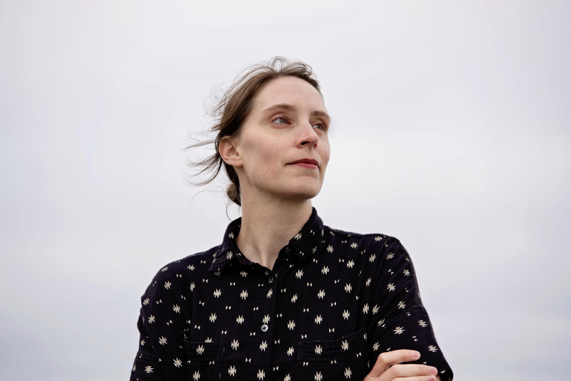 Zoë Bossiere, in a black collared button-down and wind-swept hair, looks into the distance.