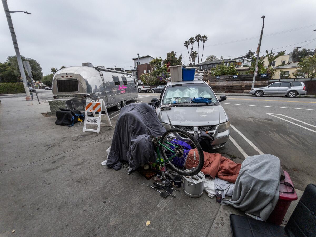 A long, silver camping trailer and a passenger car are parked along a sidewalk where someone's bedding is laid out. 