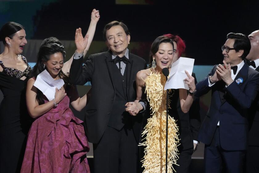 A cast of actors win black-tie attire on a stage. A woman in a gold and black gown reads a speech