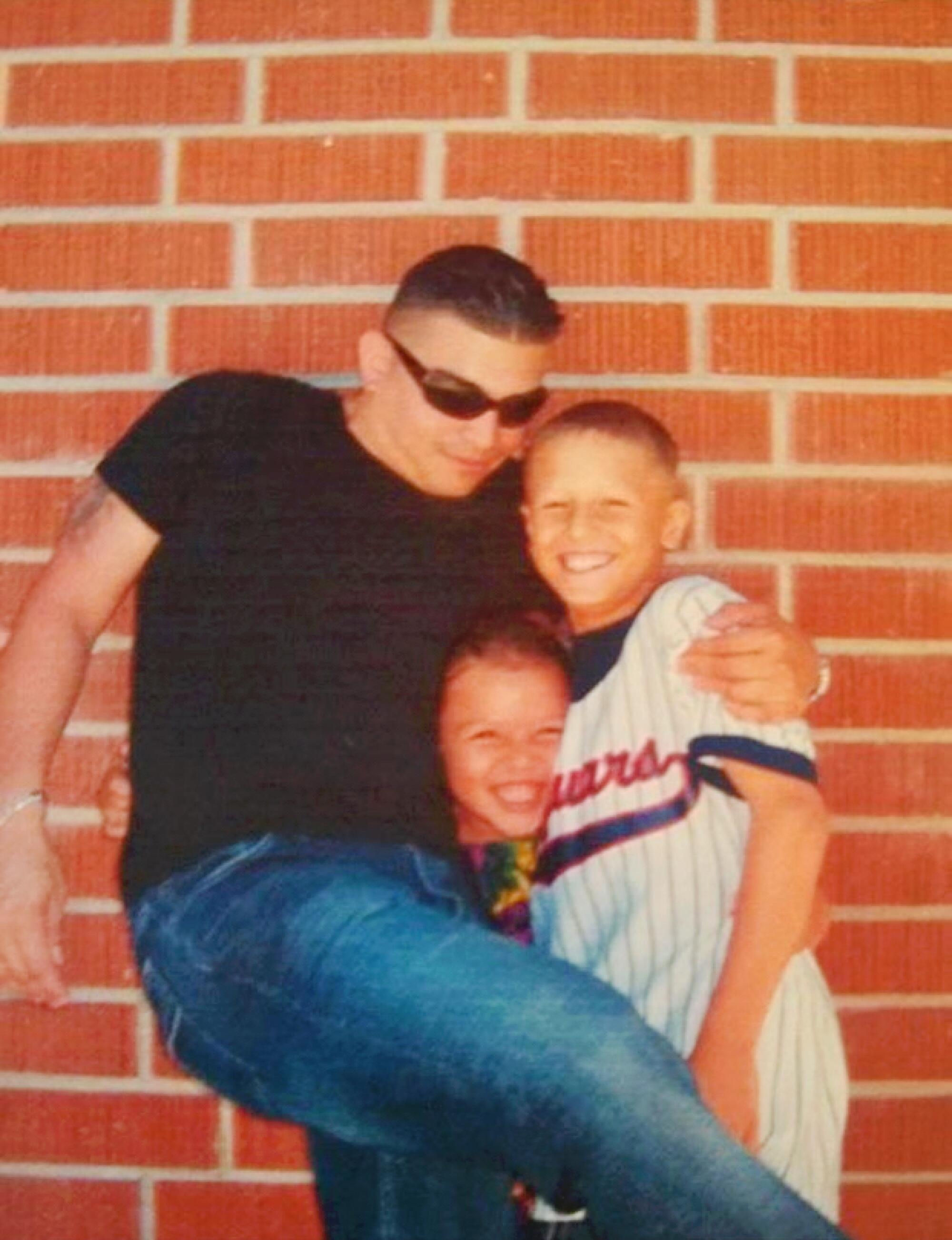 A man hugs two children with one arm in front of a brick wall.