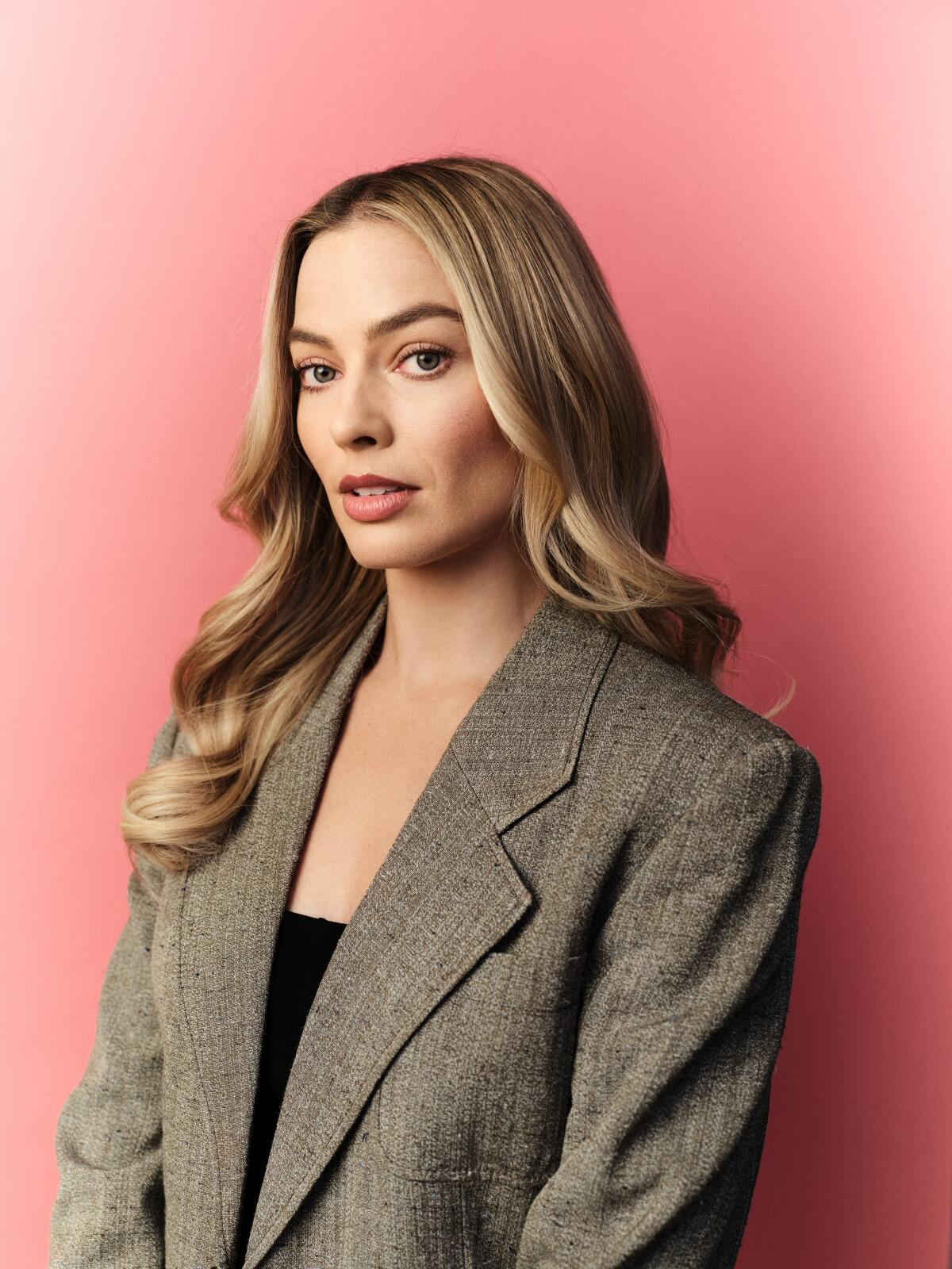 Margot Robbie in a gray oversized blazer and black top standing against a peachy, pink background