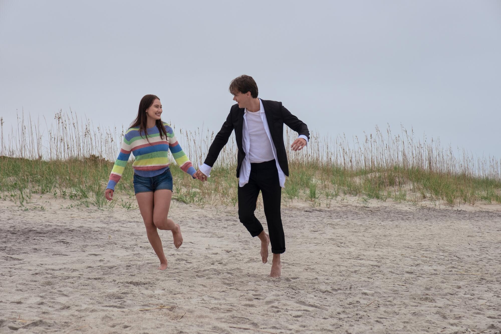A teenage girl and boy run on a beach holding hands.