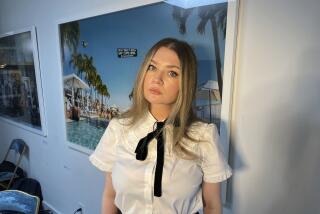 Anna Delvey in a white button down shirt, a black tie and black pants posing in front of a poster with a tropical setting