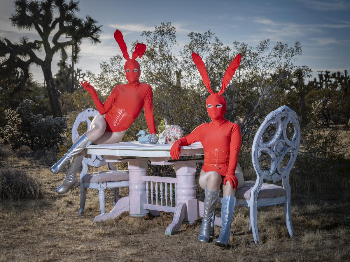 Two masked women in surreal red bunny costumes sit on patio furniture, surrounded by Joshua trees and desert scrub.