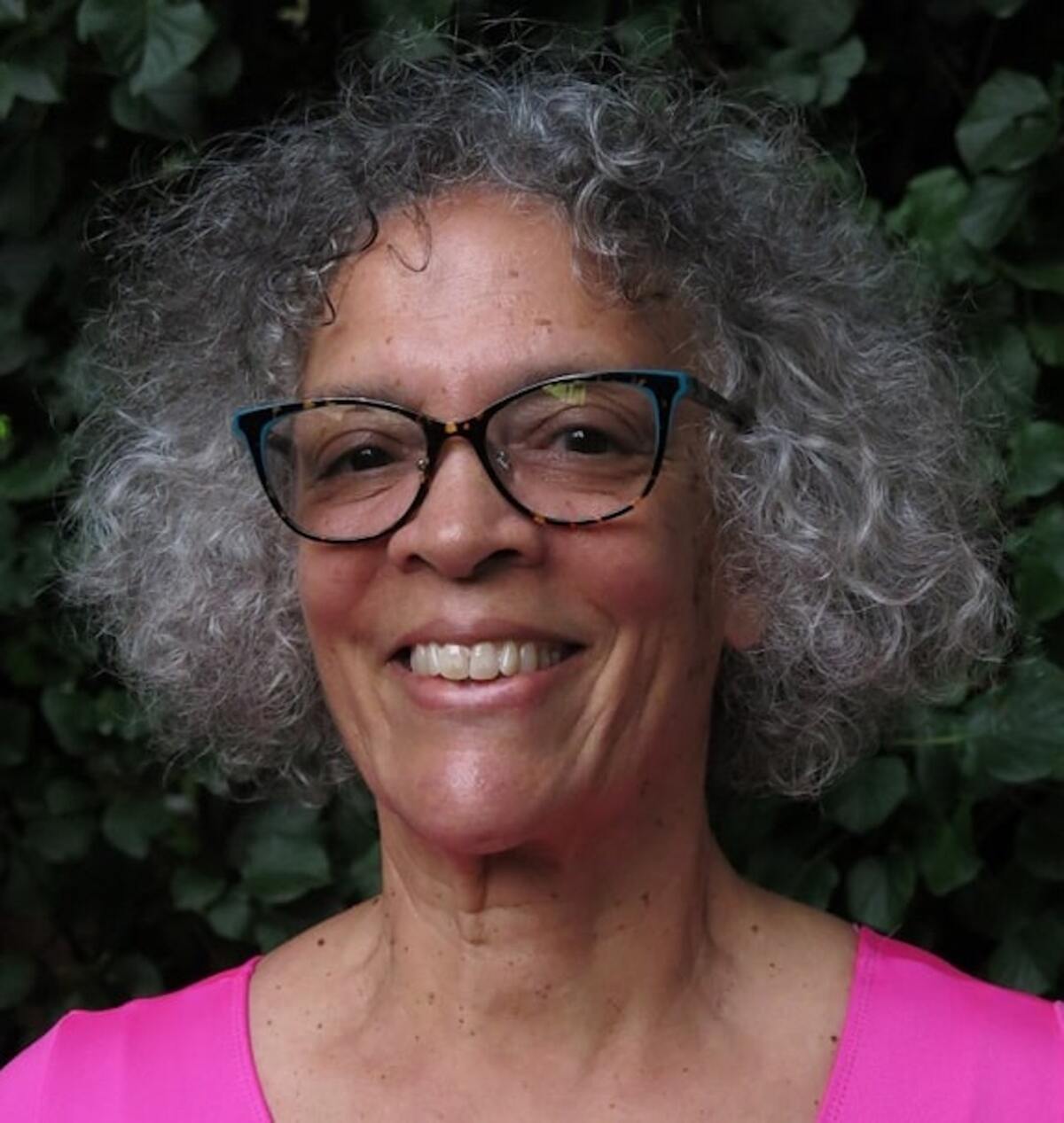 A women with gray hair, glasses and a bright pink top smiles