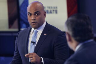 A man with a clean-shaven head, in dark suit and light blue tie, speaks while looking at a man, seen from behind