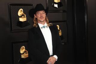 A man with long, blond hair posing in a black suit and black cowboy hat
