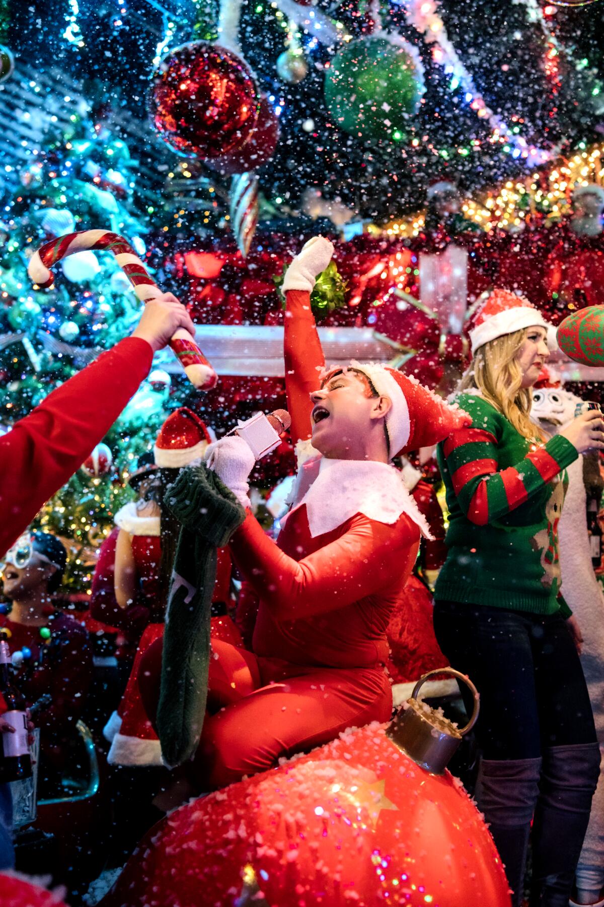 A man dressed as a Christmas elf singing atop a giant ornament at a party.