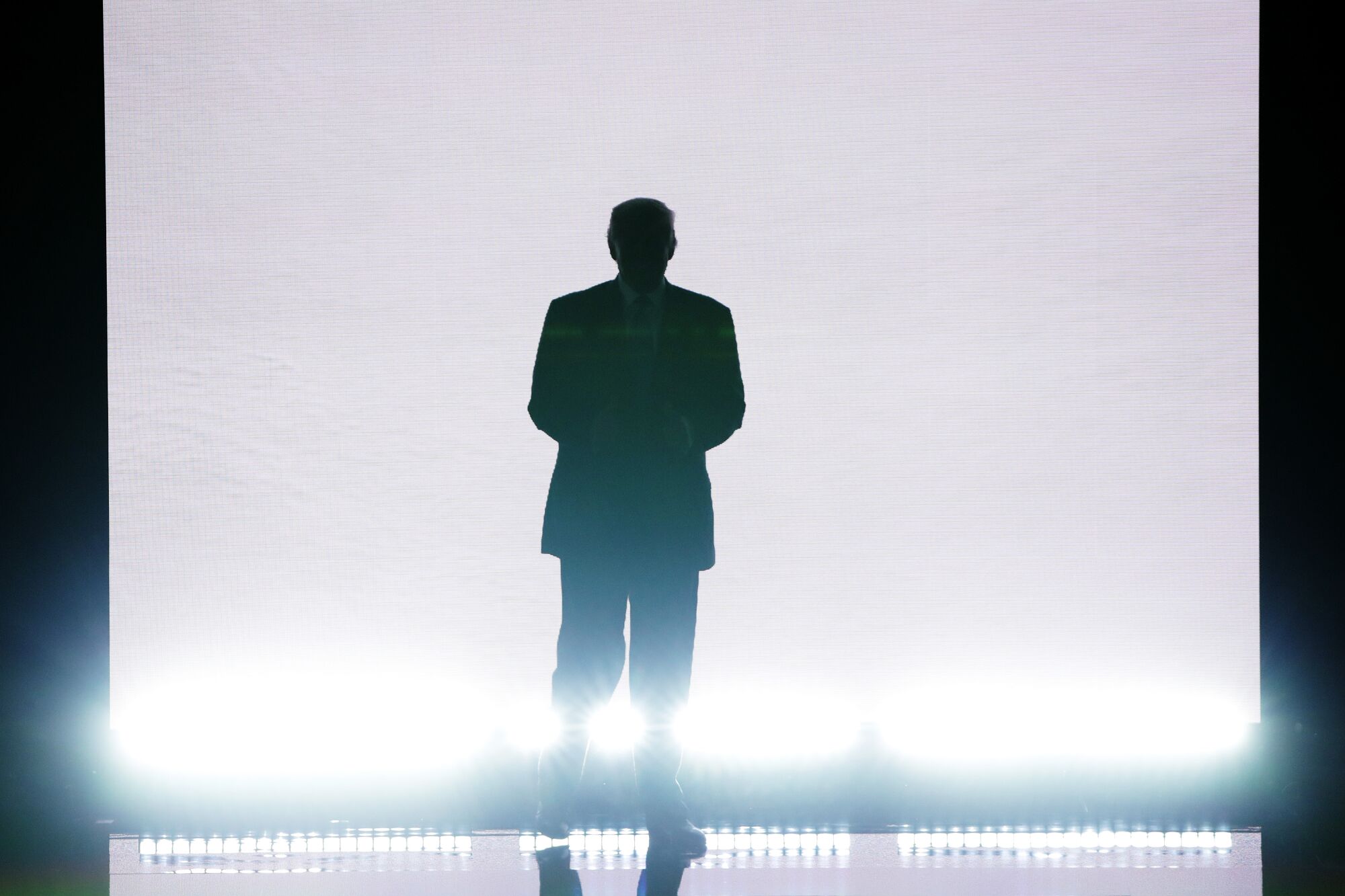 Donald Trump is lighted from behind with a blank screen behind him while walking on a stage.